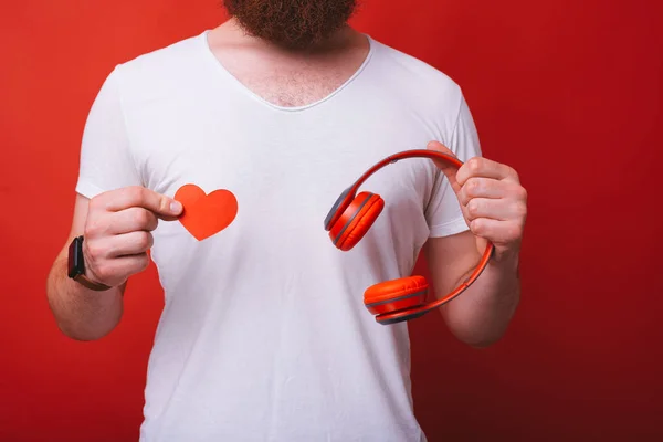 我喜欢音乐！红色主题。男子拿着一张红色的心形纸和一对红色耳机在红色背景. — 图库照片