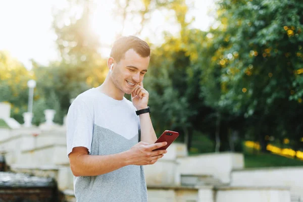 Young cheerful man is listening to the music an looking in the phone in park. — Stock Photo, Image