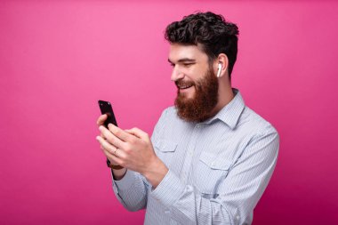 Genç sakallı adam pembe arka planda telefon ve kulak lıklarını kullanıyor. Kablosuz teknoloji.