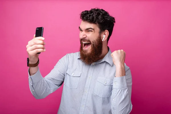 Mladý vousatý muž, který jednou rukou dělá vítězný gesto, zatímco se dívá na telefon, křičí a nosí náušník. — Stock fotografie