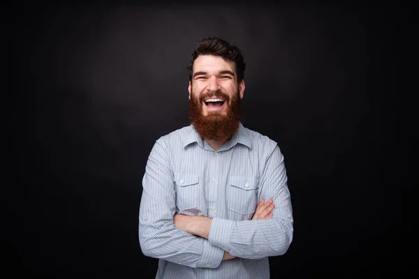 Счастье и смех. Портрет молодого бородатого мужчины, стоящего со скрещенными руками на черном фоне, смеющегося — стоковое фото