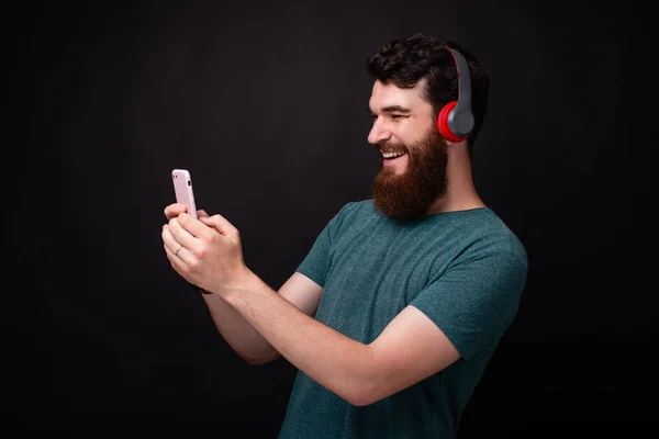 Joven barbudo haciendo una selfie y escuchando la música a través de auriculares rojos sobre fondo negro . —  Fotos de Stock