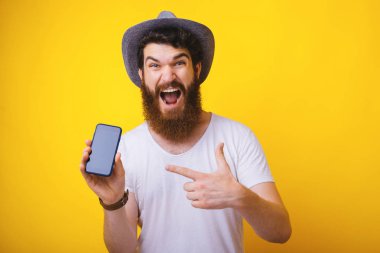 Heyecanlı sakallı adamın fotoğrafı, şapka takan, kameraya bağıran, mobil ekranı işaret ederken, sarı arka plan üzerinde duran