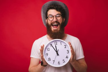 Yakışıklı heyecanlı sakallı adam fotoğrafı, bir kovboy şapkası giyen, kırmızı arka plan üzerinde duran saat tutan 