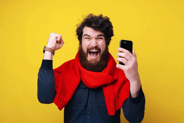 ¡Sí! Chico barbudo guapo celebrando con el brazo levantado, usando una bufanda roja, y sosteniendo teléfono inteligente sobre fondo amarillo — Foto de Stock