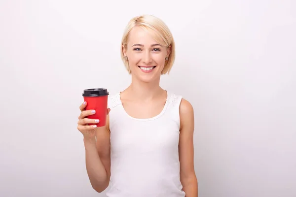 Cheerful young woman, looking at camera with toothy smile,  while holding a red cup of coffee, over isolated background — Stock Photo, Image