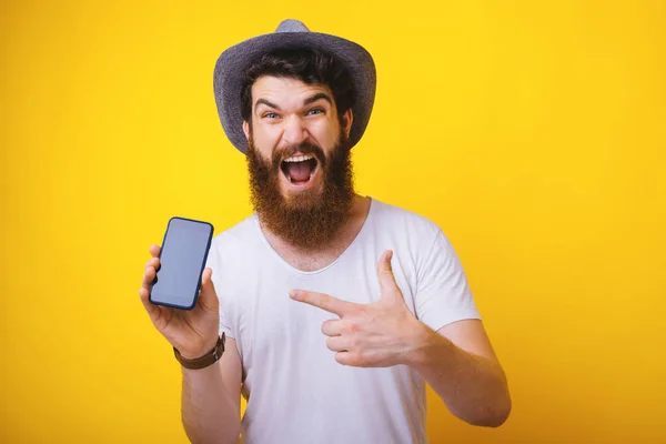 Fotó izgatott szakállas srác, fárasztó kalap, sikoltozva a kamera, miközben mutat a mobil képernyő, állva sárga háttér — Stock Fotó