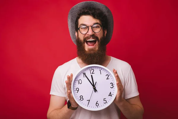 Foto de hombre barbudo guapo emocionado, con un sombrero de vaquero, sosteniendo el reloj de pie sobre el fondo rojo — Foto de Stock