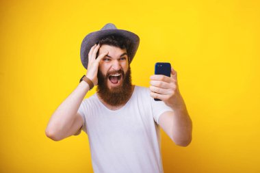 Yakışıklı sakallı adam Fotoğraf, cep telefonu tutan ve sreen hayran görünümlü, yelow arka plan üzerinde 