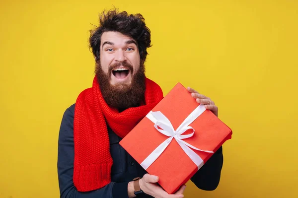 Foto de cara barbudo, vestindo lenço vermelho, olhando animado para a câmera enquanto segurando uma caixa de presente e em pé sobre fundo amarelo — Fotografia de Stock