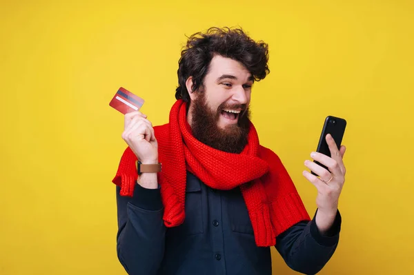 Tipo barbudo emocionado, usando el teléfono móvil y la tarjeta de crédito, compras en línea, de pie sobre fondo aislado — Foto de Stock