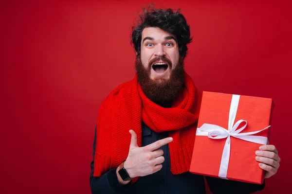 Excited bearded guy, wearing red scarf pointing at gift box, with amazed face and openeed face