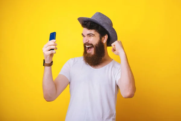 Foto de un guapo barbudo con sombrero, sosteniendo un teléfono móvil y celebrando con los brazos arriba sobre fondo amarillo — Foto de Stock
