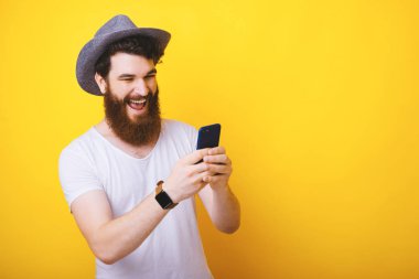 Yakışıklı sakallı adam fotoğrafı, bir şapka giyiyor, mutlu telefonda bir metin mesajı yazarak, sarı arka plan üzerinde 