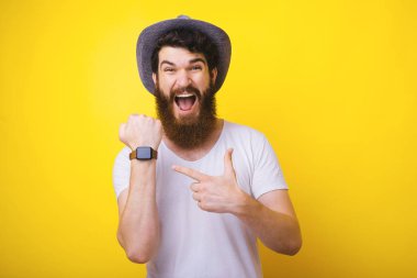 Kovboy şapkalı neşeli sakallı bir adam, heyecanla kol onun smartwatch işaret, izole sarı bir arka plan üzerinde 