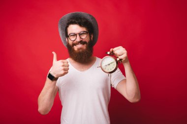 Şapka ve gözlük gülümseyen ozan adam Portresi, bir vintage saat tutan ve kırmızı arka plan üzerinde başparmak gösteren  