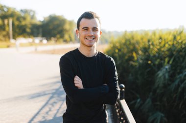 Spor konsepti, spor giyimli genç adam, korsize elleri ile, gülümseyen ve kamera poz, parkta sabah koşmak