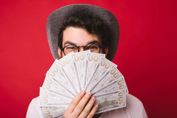 Photo of handsomea guy wearing hat and glasses, covering his face with a lot of money, over red background — Stock Photo, Image