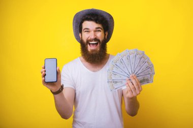 Sakallı adam, şapka takıyor, elinde çok para tutuyor ve mobil ekranı gösteriyor.