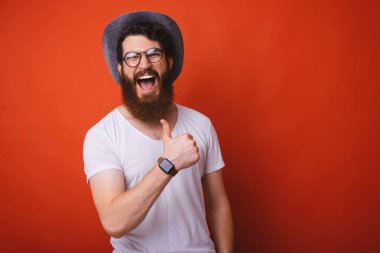 Şapka ve gözlük heyecanlı sakallı adam fotoğrafı, kırmızı backgroung üzerinde başparmak gösteren 