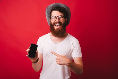 Bir şapka ve gözlük takan neşeli sakallı adam, kırmızı arka plan üzerinde cep telefonu işaret 
