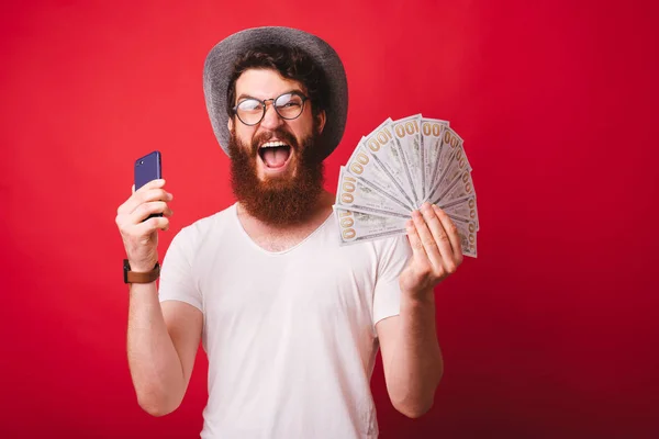 Hombre hipster barbudo asombrado sosteniendo teléfono inteligente y dinero en efectivo de pie —  Fotos de Stock