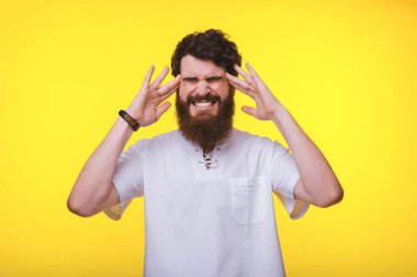Sakallı bir adam sarı bir duvarın yanında baş ağrısı olduğunu gösteriyor.