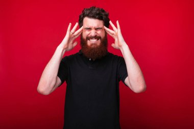 Genç sakallı adam kırmızı arka plan yakınında baş ağrısı yaşıyor.