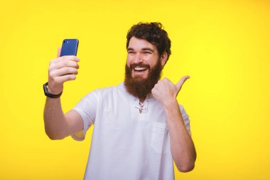 Neşeli sakallı ve genç adam başparmak ap ve sarı bir duvara yakın bir gülümseme ile selfie alıyor.