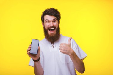 Çıkan sakallı adam bir akıllı telefon tutuyor ve sarı duvarın yanında başparmağını gösteriyor..