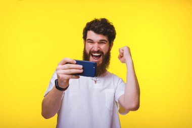 Neşeli genç sakallı adam akıllı telefon oynuyor ve sarı backround üzerinde yükseltilmiş yumruk ile zaferini kutluyor.