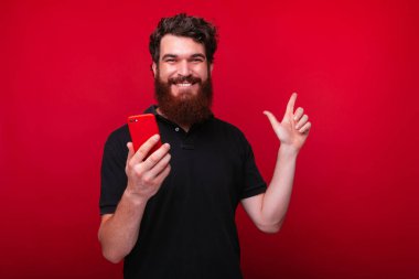 Neşeli genç sakallı adam bir akıllı telefon tutuyor ve kırmızı arka plan üzerinde işaret. 