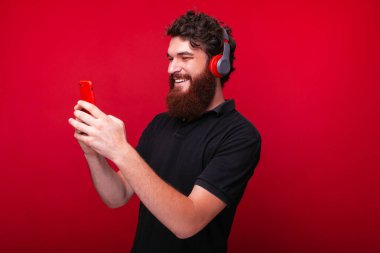 Neşeli genç adam kırmızı bir arka plan üzerinde kafasına kulaklık ile telefon bakıyor.
