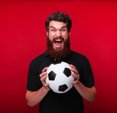 Genç sakallı adam çığlık atıyor ve kırmızı arka planda bir futbol topu tutuyor