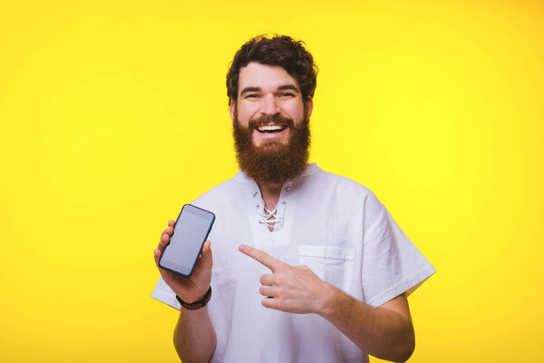 Cheerful bearded man is pointing to a smartphone near yellow background.