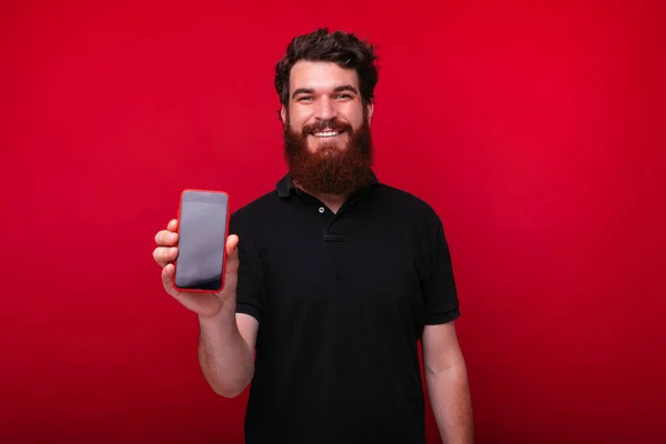 Barbudo hombre está sonriendo a la cámara y mostrando un teléfono inteligente . — Foto de Stock