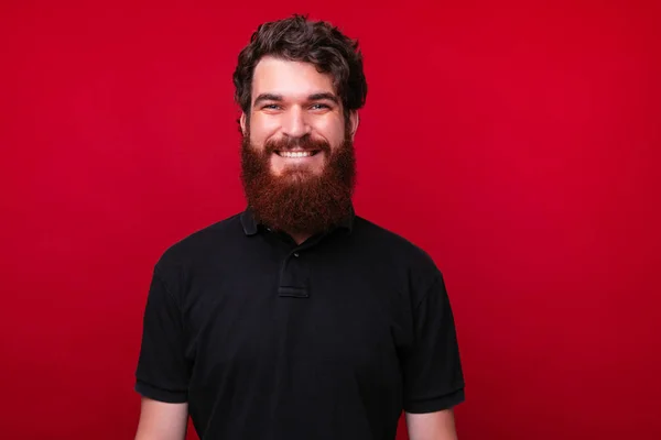 Joven barbudo alegre está mirando a la cámara sobre un fondo rojo . —  Fotos de Stock