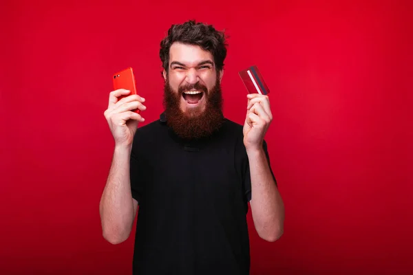 Joven barbudo alegre está gritando una celebración de un teléfono inteligente y una tarjeta de crédito sobre fondo rojo . — Foto de Stock