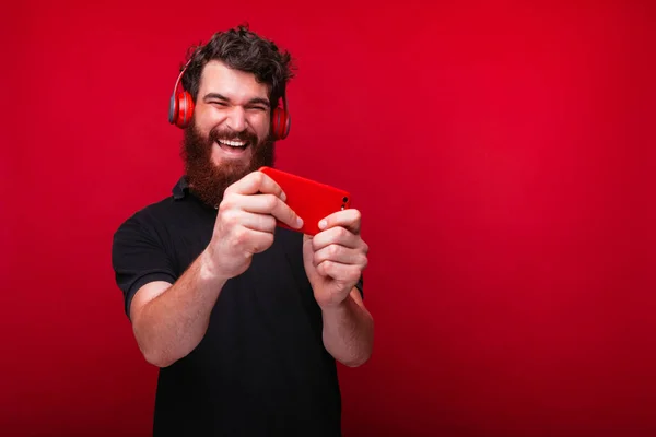 Joven barbudo alegre está escuchando la música y jugando en el teléfono inteligente sobre fondo rojo . —  Fotos de Stock