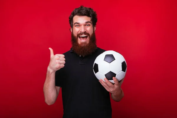 Wesoły Brodaty mężczyzna pokazuje kciuka i trzyma piłkę nożną na czerwonym tle. — Zdjęcie stockowe