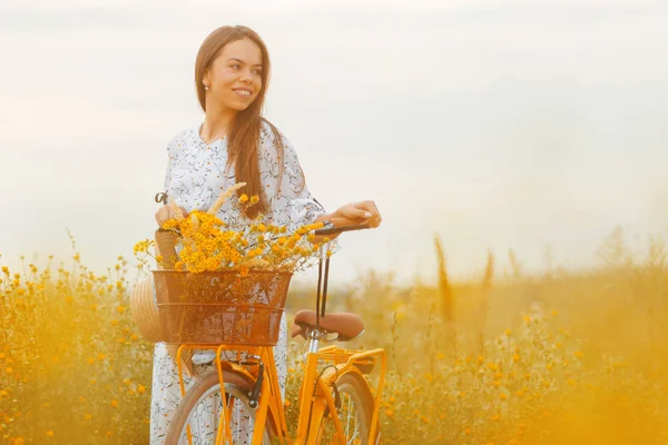 Retrato de una hermosa mujer atractiva sosteniendo una bicicleta amarilla retro y mirando en algún lugar mientras sonríe — Foto de Stock