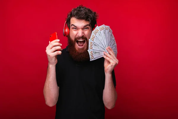 Excited bearded guy,with headphones on head, holding mobile phon — Stock Photo, Image