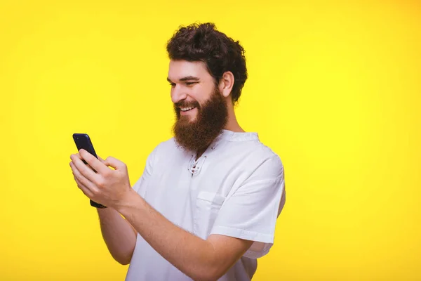 Joven barbudo escribiendo o tocando algo en su teléfono inteligente sobre fondo amarillo . —  Fotos de Stock