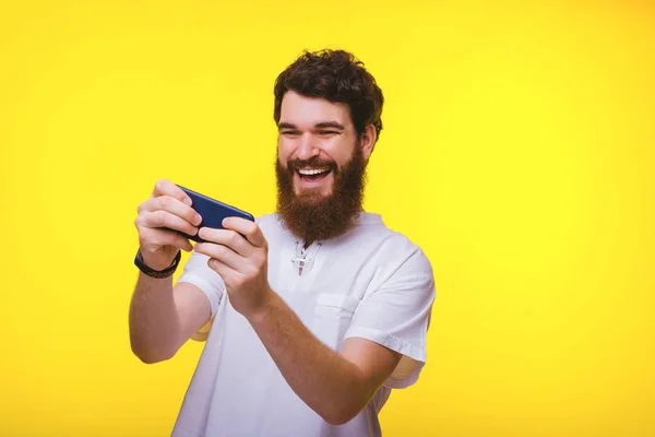 Alegre hipster barbudo jugando en sus juegos de carreras de teléfonos inteligentes sobre fondo amarillo. Diversión en verano . —  Fotos de Stock
