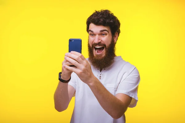 Hombre barbudo haciendo una cara excitada mientras mira su teléfono inteligente en el fondo amarillo . —  Fotos de Stock