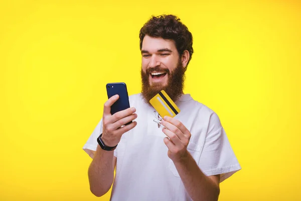Feliz barbudo hipster está mirando su teléfono y la celebración de una tarjeta de crédito . —  Fotos de Stock