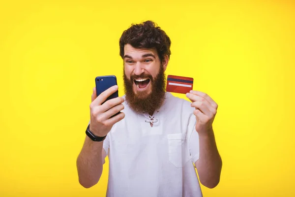 震惊的胡子的家伙看着他的手机，而拿着他的红色信用卡在黄色背景. — 图库照片