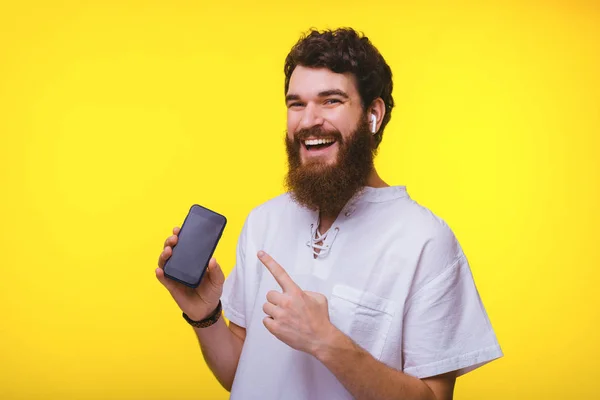 Joven barbudo alegre señalando su teléfono y escuchando música en airpods sobre fondo amarillo . —  Fotos de Stock