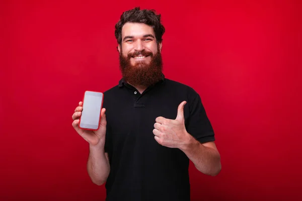 Retrato del hombre sonriente con barba mostrando la pantalla del teléfono inteligente y el gesto hacia arriba pulgares —  Fotos de Stock