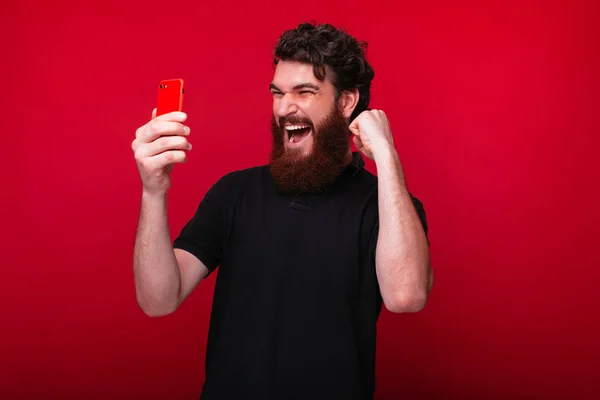 Foto di eccitato giovane uomo barbuto guardando smartphone e celebrando il successo — Foto Stock